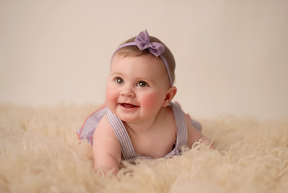 Five-month-old baby girl smiling during a milestone photoshoot, lying on her tummy on a soft cream flokati rug, wearing a lavender outfit and matching bow headband, capturing her growing personality and developmental milestone.