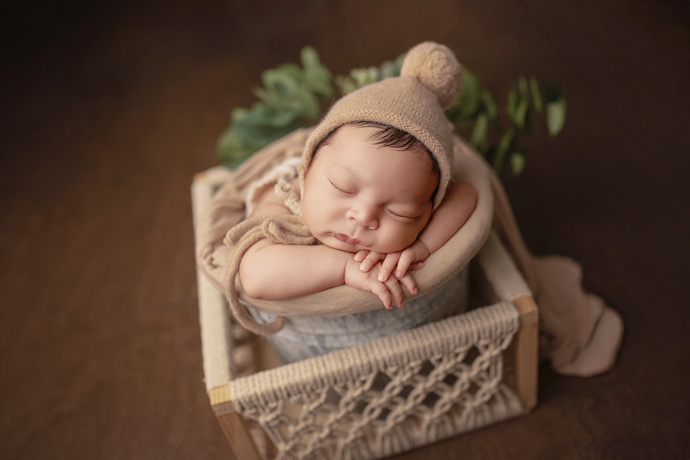 Sleeping newborn baby boy posed in a cozy bucket with his hands gently folded under his chin, wearing a neutral knit bonnet during a professional newborn photography session in Conroe, TX.